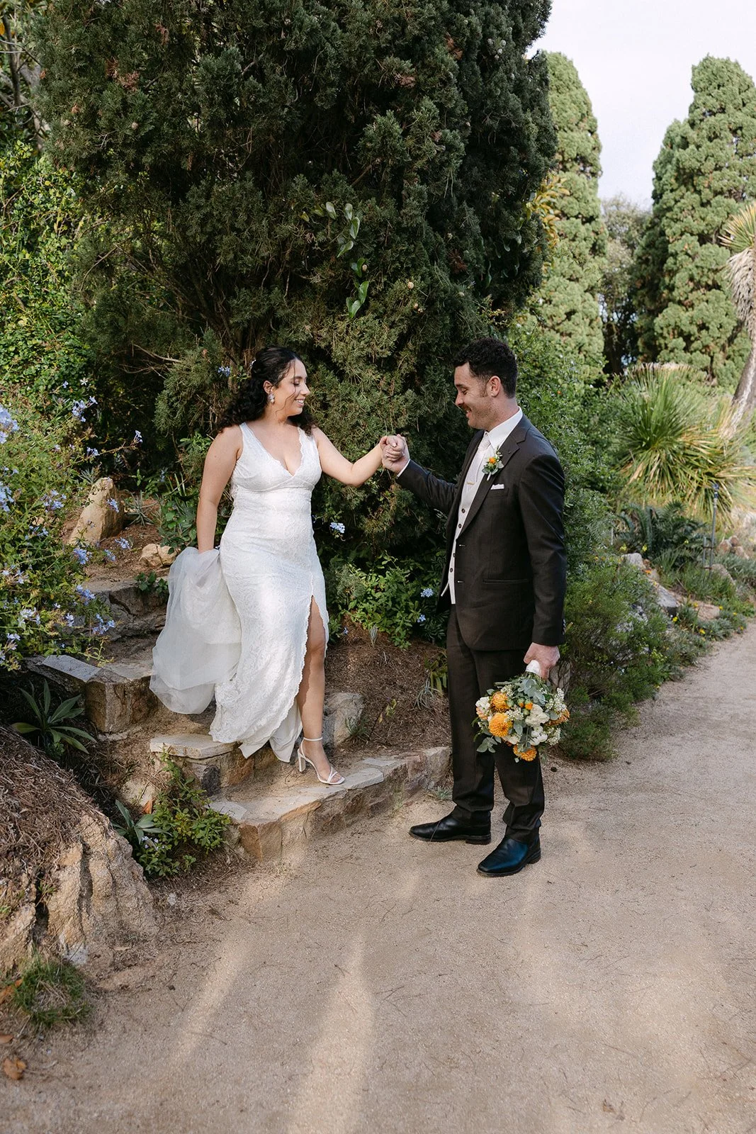 A bride and groom smiling and holding hands outdoors in a garden setting, with the groom holding a bouquet of flowers.