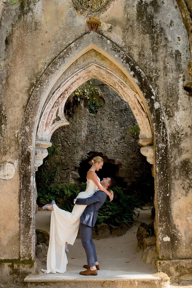 A couple dressed in wedding attire, with the man in a gray suit and the woman in a white wedding dress, in a romantic pose beneath an old stone archway surrounded by greenery.