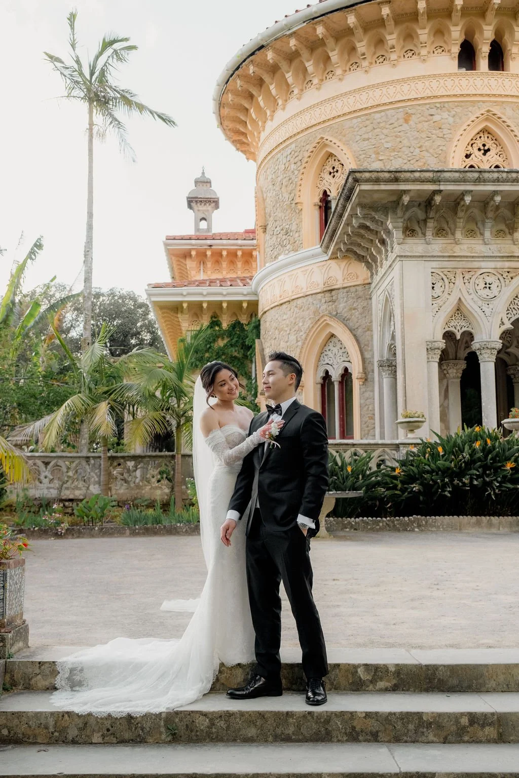 A bride in a white wedding gown and a groom in a black tuxedo pose in front of a historic castle with intricate architecture, surrounded by lush greenery.