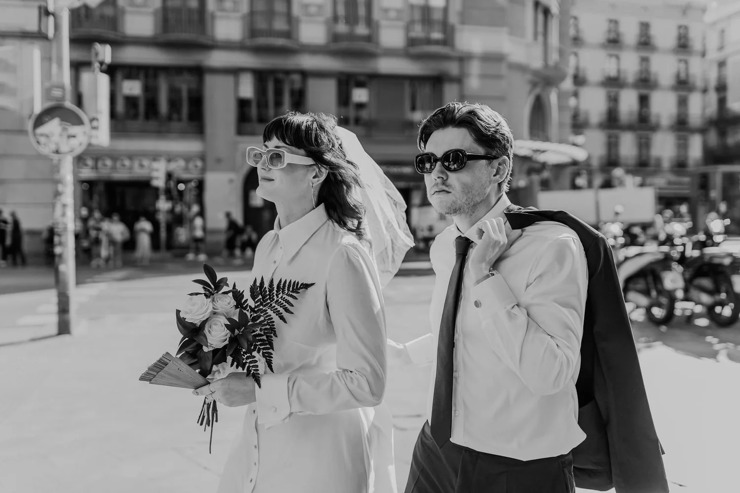 A black and white photo of a bride and groom walking outdoors. The bride is holding a bouquet and looking to the left, wearing large glasses and earrings. The groom, wearing sunglasses and a suit, walks beside her with a backpack over his shoulder.