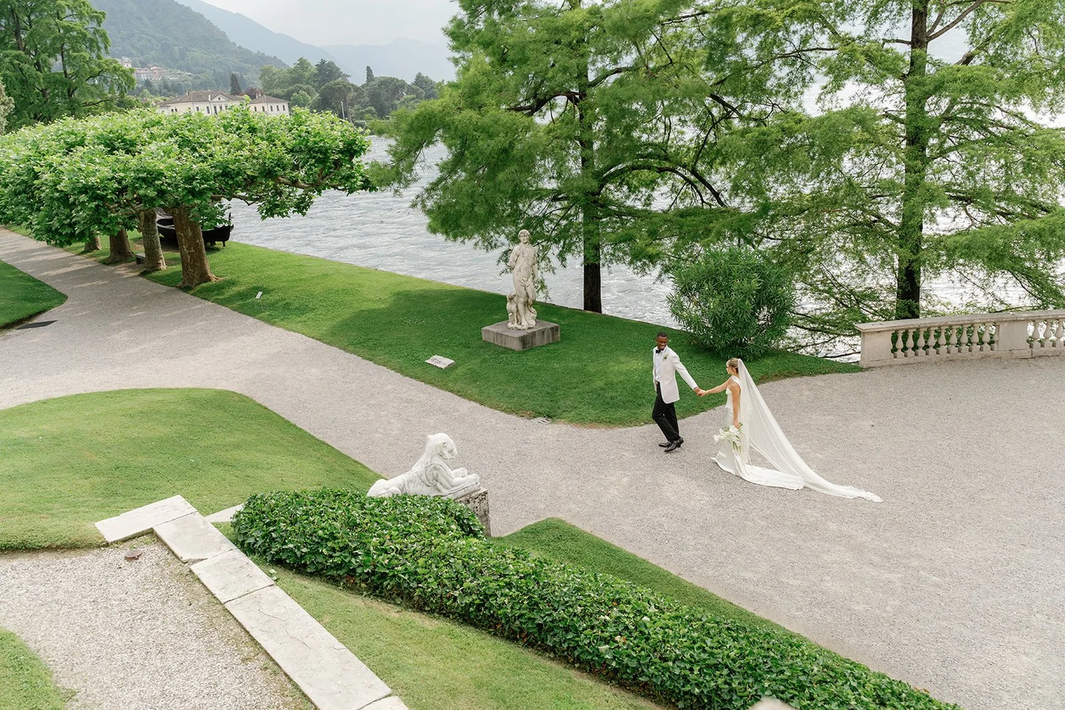 A bride and groom holding hands and walking in a lush garden near a river, with classical sculptures and large green trees.