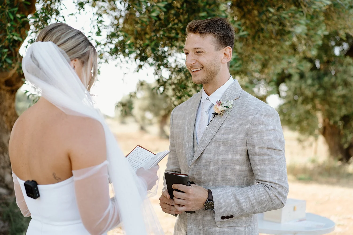A groom in a light gray suit smiling at a bride during a wedding ceremony outdoors with trees in the background.
