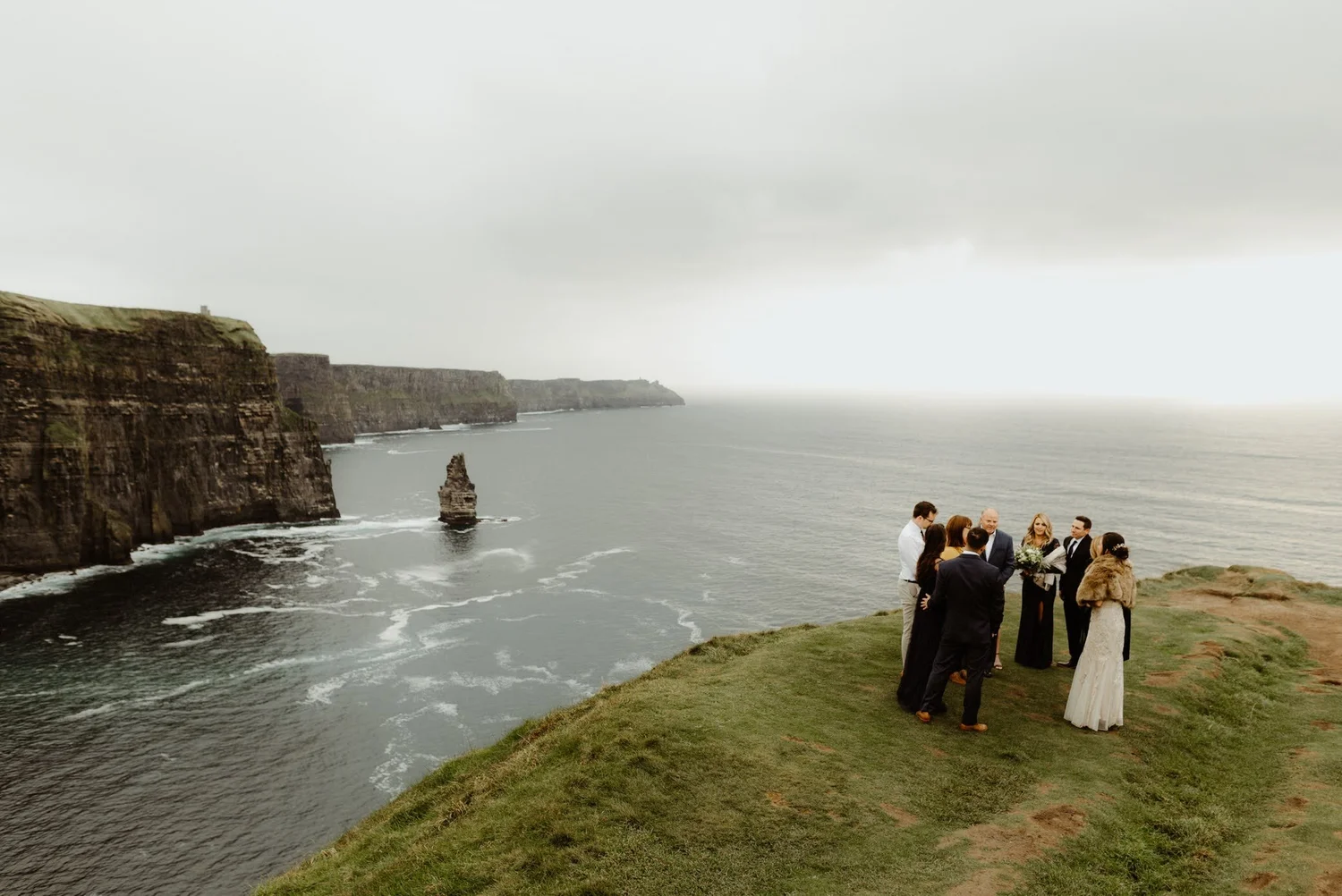 A group of people dressed in formal attire standing on a grassy cliff overlooking the ocean during an overcast day, with cliffs in the background.