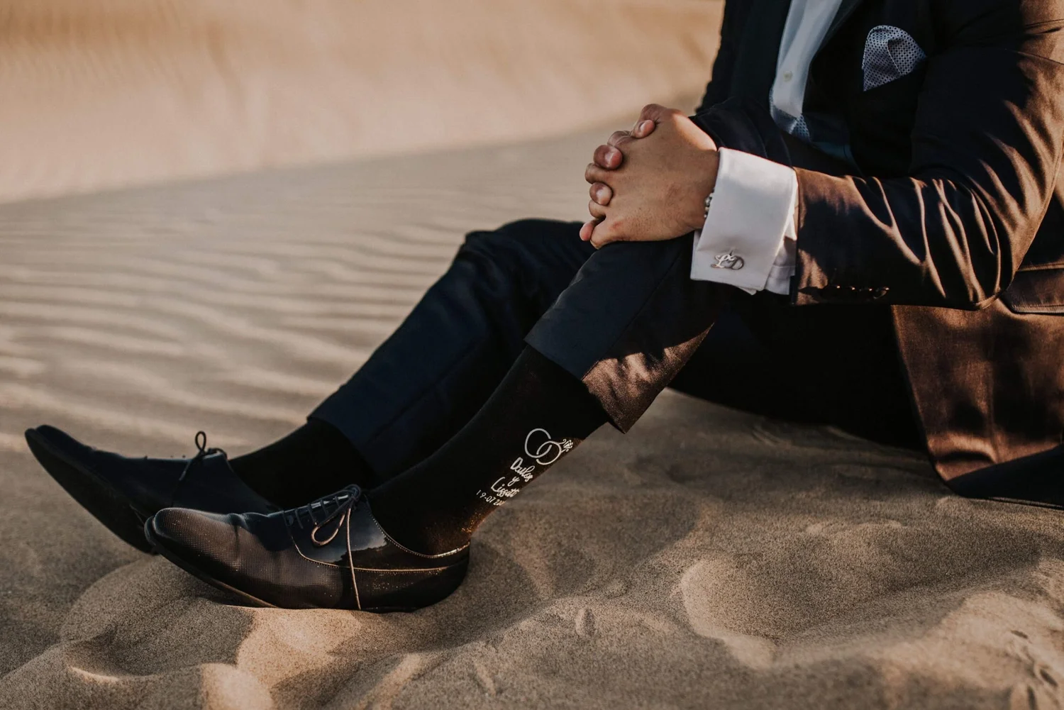 A person dressed in formal attire sitting on a sandy surface, with hands clasped and legs outstretched, wearing black shoes and black socks with writing on them.