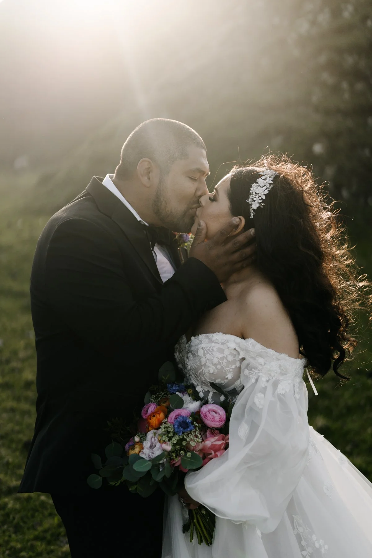 A bride and groom kissing outdoors during sunset, with the groom in a black tuxedo and the bride in a white lace wedding dress, holding a colorful bouquet of flowers.