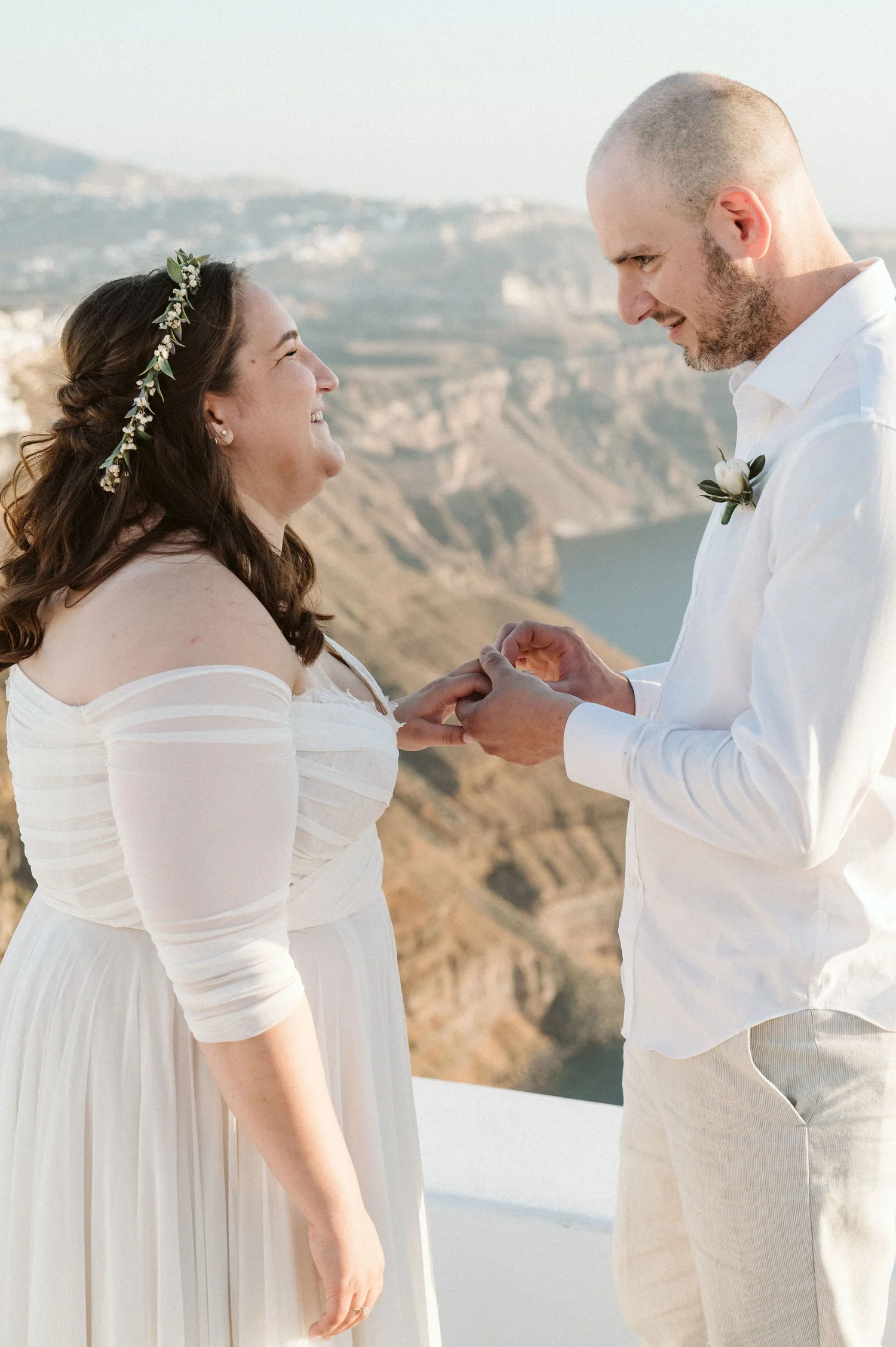 A couple at their wedding holding hands and smiling at each other, with a scenic landscape of cliffs and water in the background.