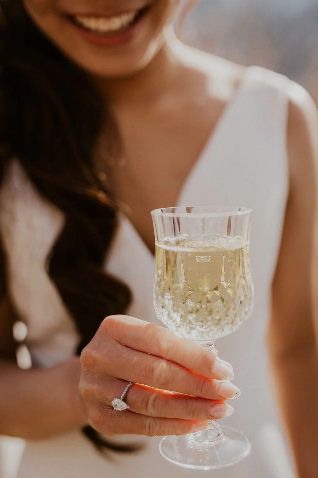 A woman in a white dress smiling and holding a champagne flute filled with bubbly champagne.