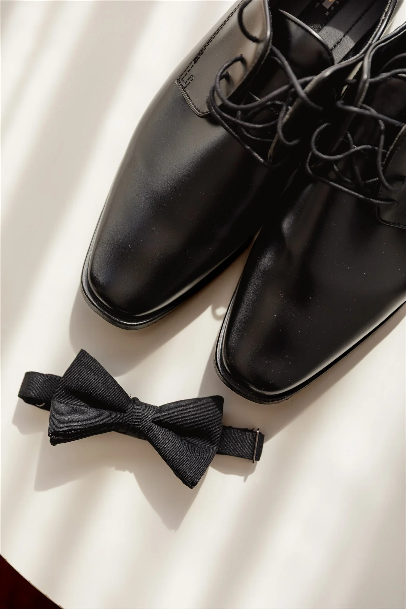 A pair of black leather dress shoes, a black bow tie, and a black strap are arranged on a white surface.