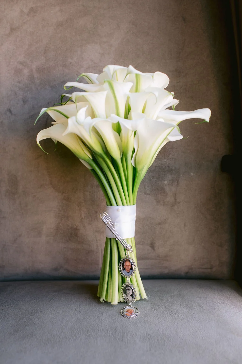 A bouquet of white calla lilies wrapped with white ribbon and decorated with small photo jewelry charms, placed on a gray surface against a gray background.