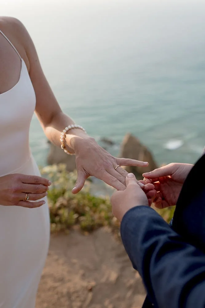 Close-up of a bride and groom exchanging rings during a beach wedding, with the ocean in the background.