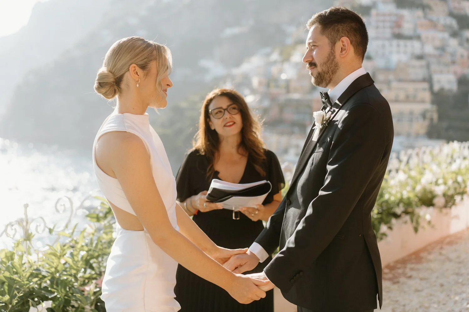 A couple dressed in wedding attire holding hands and facing each other during a wedding ceremony outdoors, with a cityscape and water in the background. An officiant stands behind them, holding a folder.