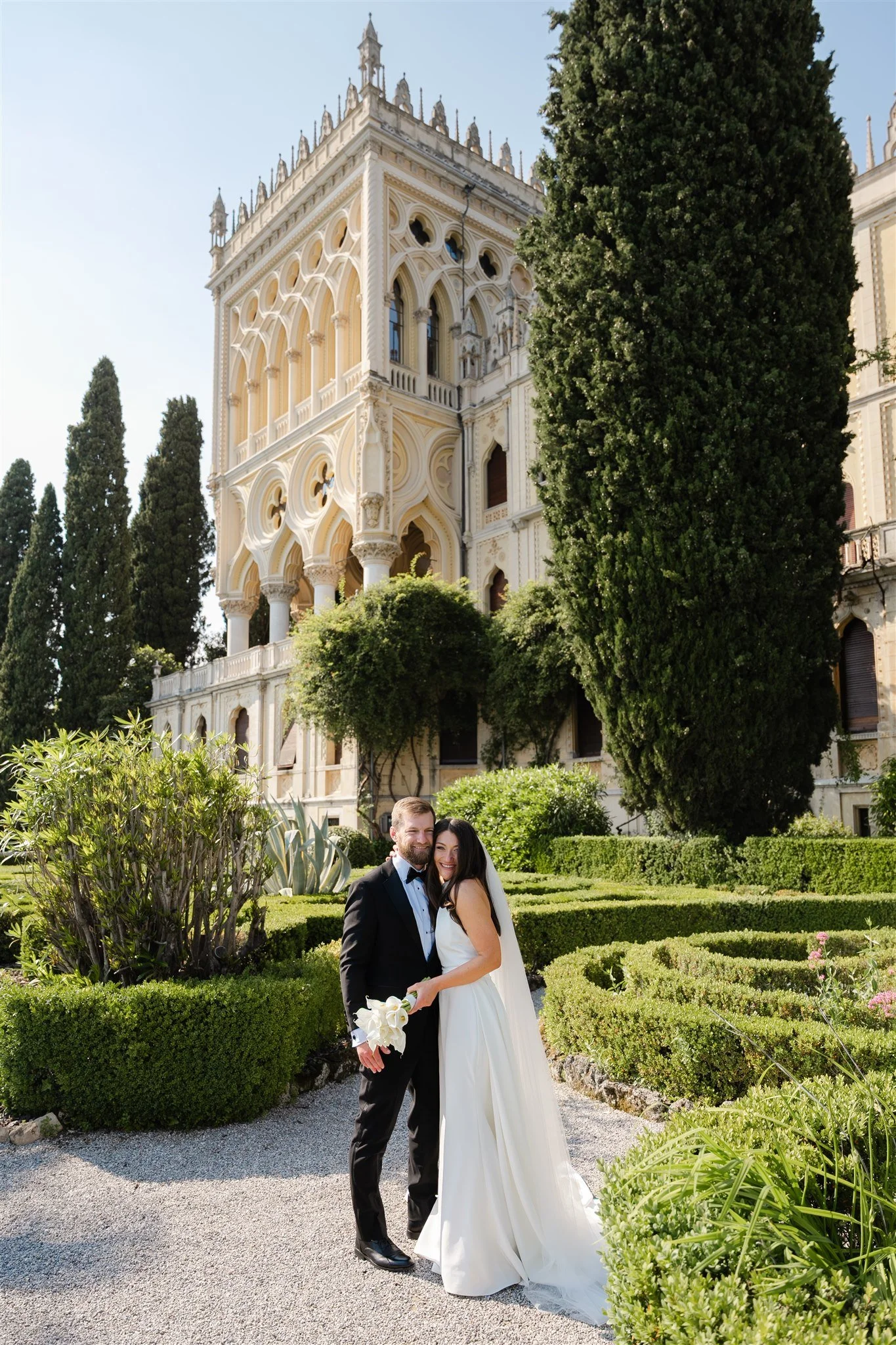 A bride and groom in wedding attire standing in a landscaped garden with tall trees and historical architecture in the background.
