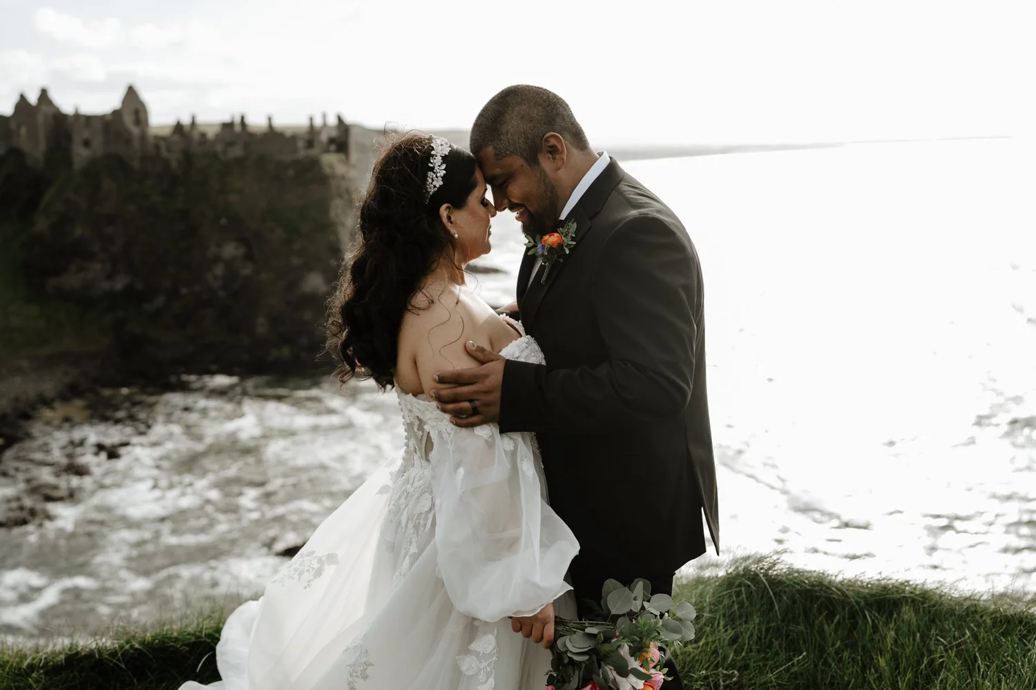 A bride and groom embrace on a beach, with a castle in the background. The bride wears a white wedding gown and headband, and the groom wears a black suit with a boutonniere. They are smiling with their foreheads touching.