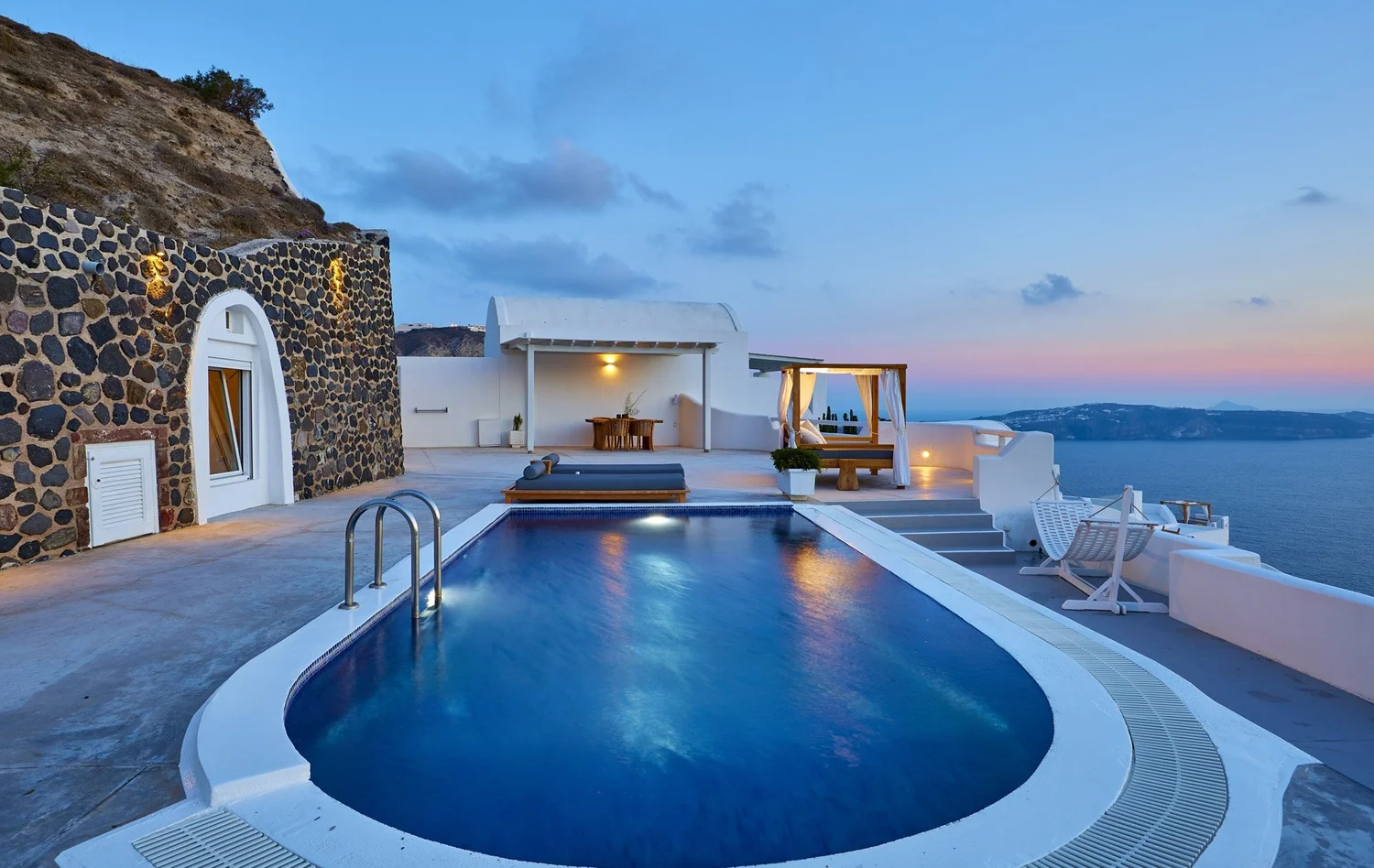 Luxury outdoor pool area on a hillside with white and stone buildings overlooking a body of water at dusk. The pool is lit, with seating and lounge areas nearby, and the sky is a soft blue with scattered clouds.