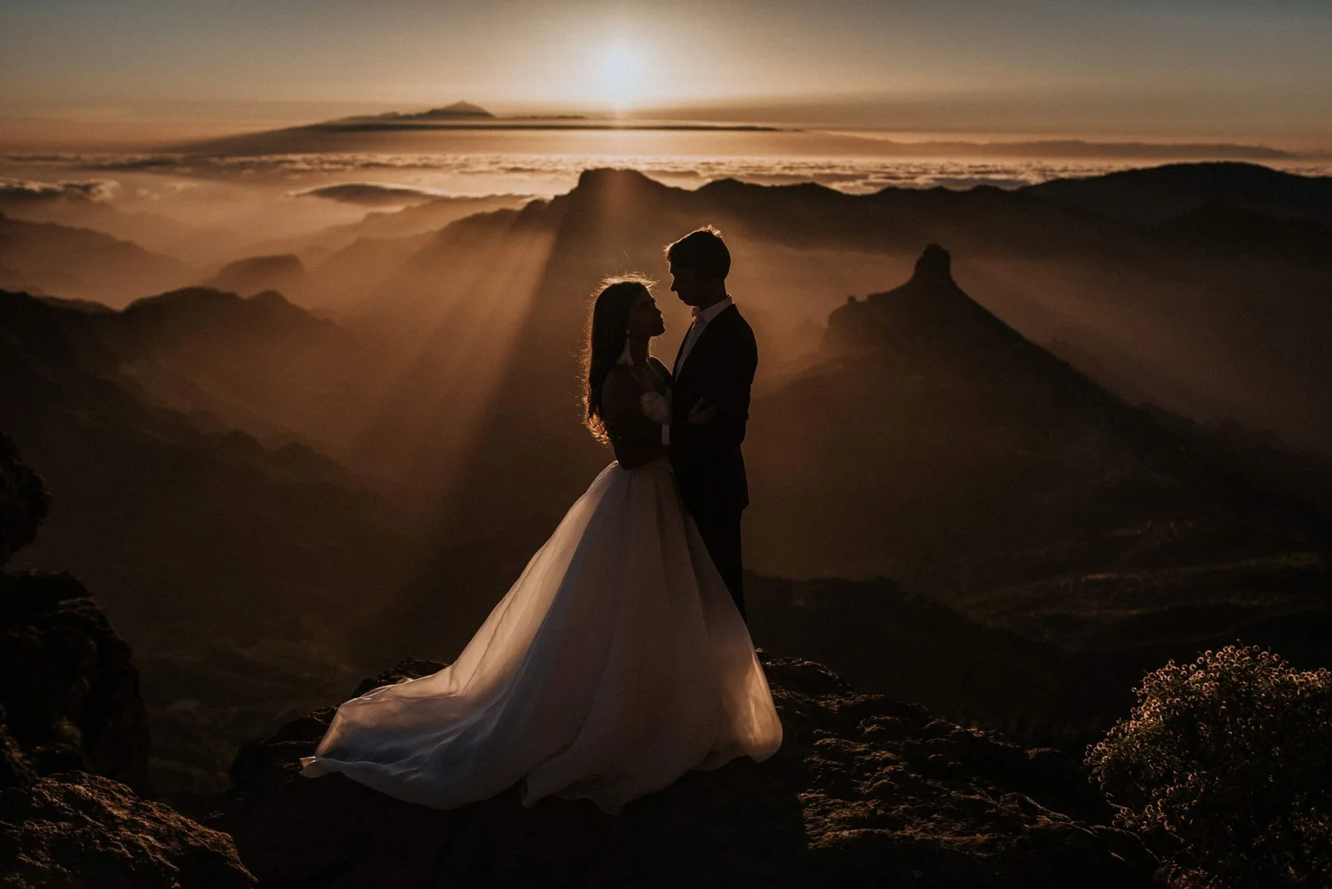 A silhouette of a couple dressed in wedding attire standing on rocky terrain during sunset with mountains and a cloudy sky in the background.