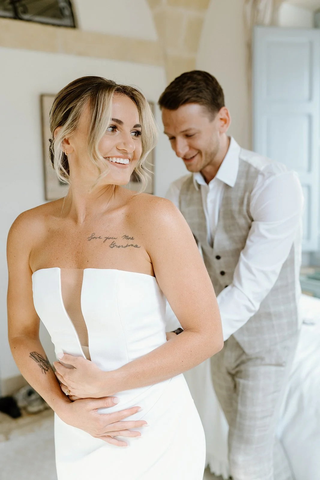 A woman in a white strapless dress is smiling while a man in a light-colored vest and pants helps her get ready.