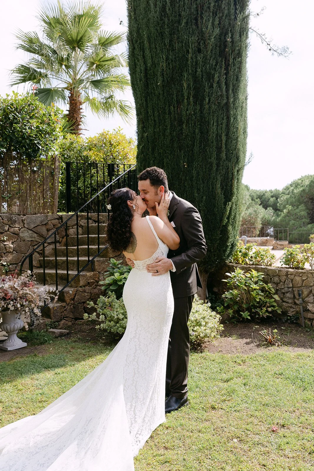 A bride and groom kiss outdoors in a garden, surrounded by greenery and tall trees, with stone steps and a metal railing in the background.