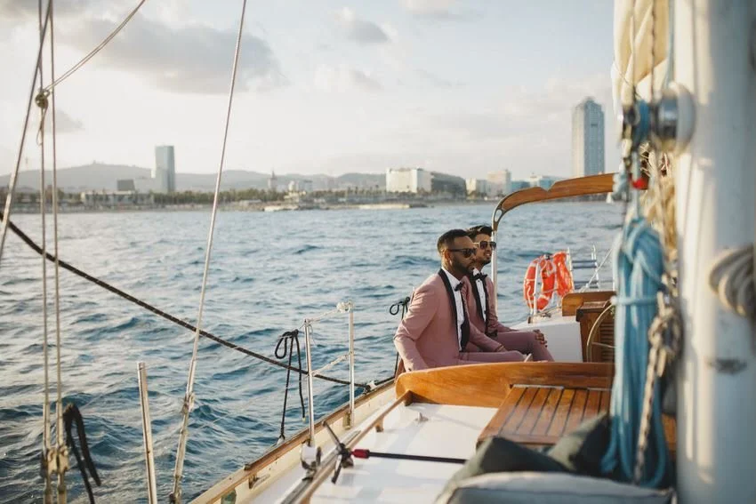 Three men dressed in suits, sitting on a sailboat, with a city skyline and water in the background.