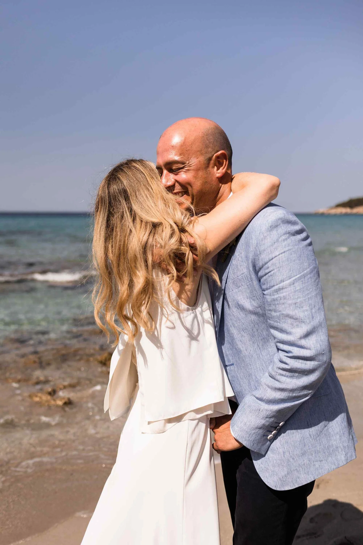 A couple dressed in formal attire hugging on a beach with the ocean and a distant coastline in the background.