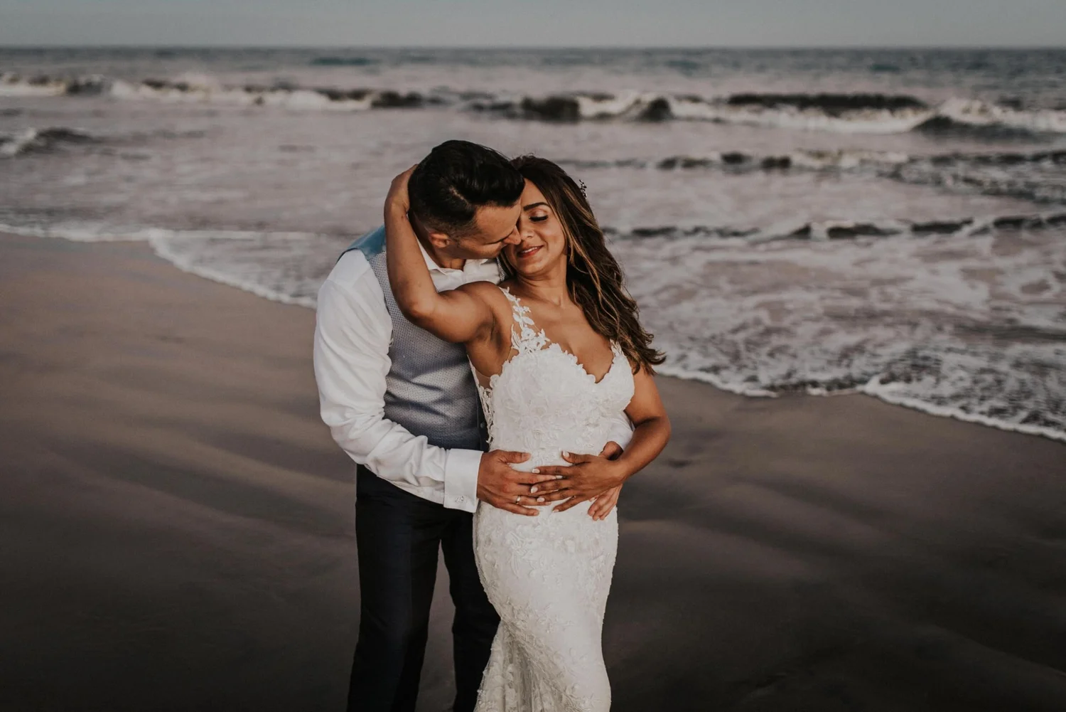 A couple embracing on the beach, the man kissing the woman's forehead, she is wearing a white lace wedding dress, and the man is in a white shirt with suspenders, with ocean waves in the background.