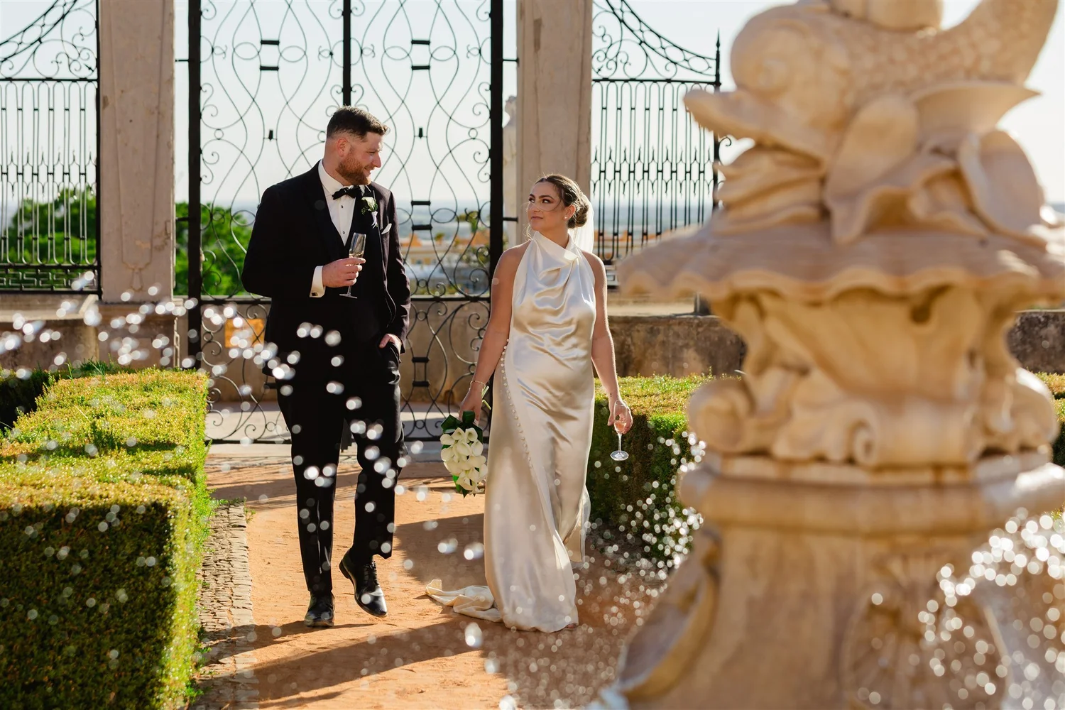 A bride and groom walking together outdoors near an ornate fountain, holding drinks, with decorative iron gates in the background, during a wedding celebration.