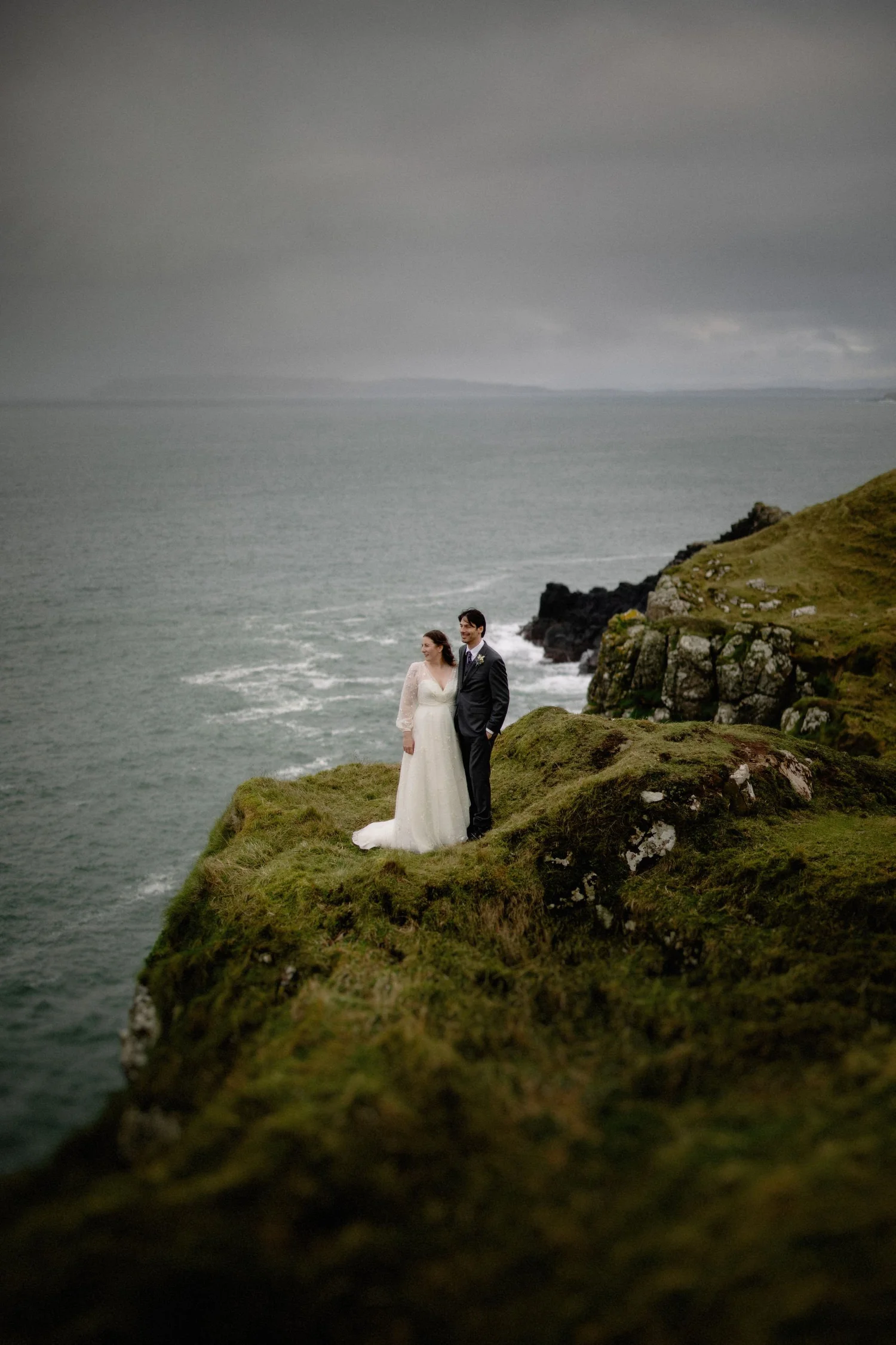 A bride and groom standing together on a grassy cliff overlooking the ocean.