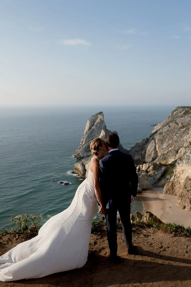 A bride and groom standing on a cliff overlooking the ocean, sharing a kiss during their wedding.