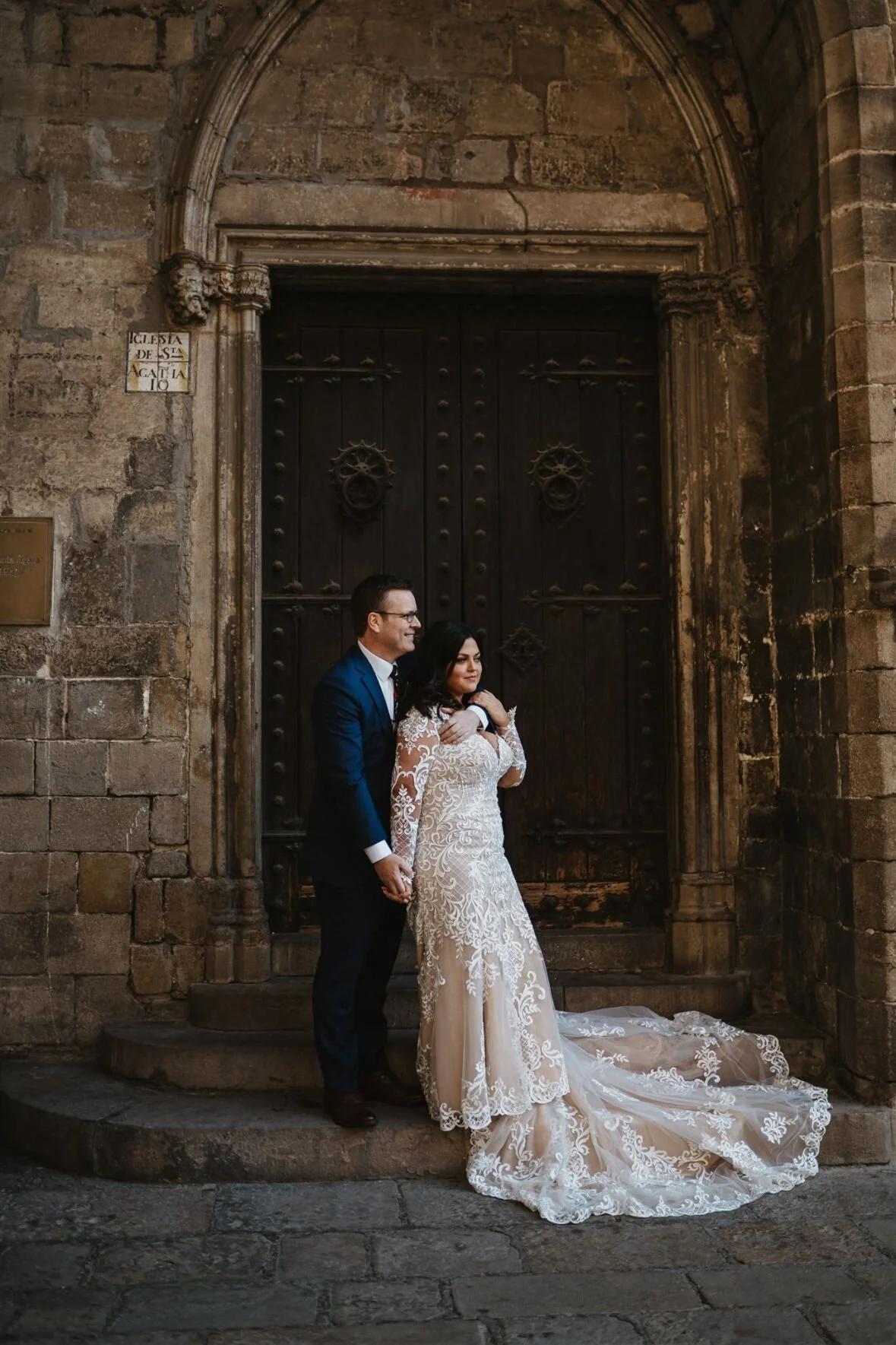 A couple in wedding attire standing in front of an old wooden door with stone walls, holding hands and looking at each other. The bride is wearing a white lace wedding gown with a long train, and the groom is in a dark suit.