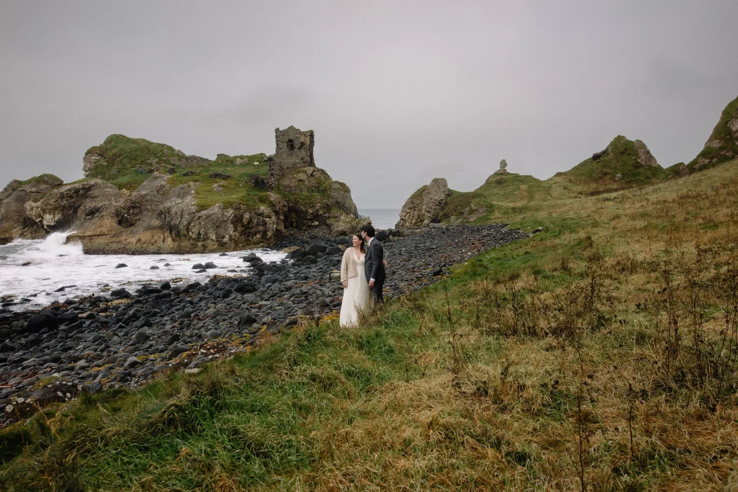 A couple in wedding attire standing on a rocky, grassy beach with a historic stone tower and rugged cliffs in the background under an overcast sky.