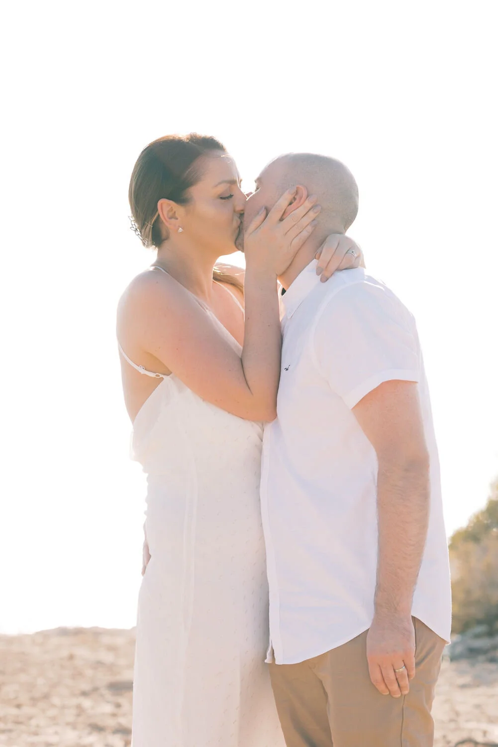 A couple sharing a kiss outdoors, with bright sunlight behind them, overlooking a beach or sandy area.