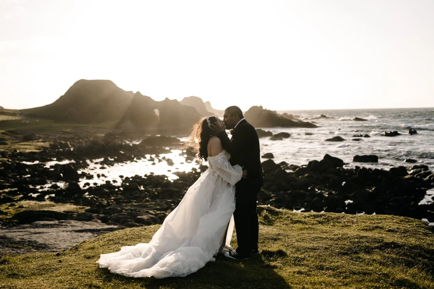 Couple in wedding attire sharing a kiss on a rocky beach with a large hill and ocean in the background at sunset.