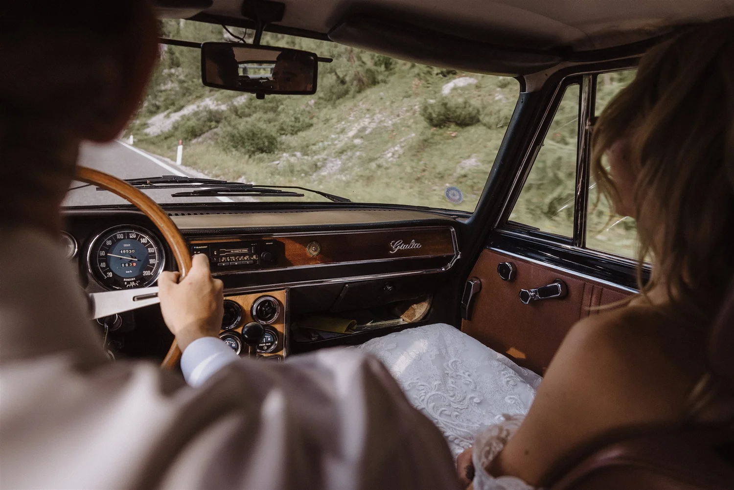 People riding in a vintage car on a mountain road, with a woman in a white dress and a man driving, uphill landscape visible through the windows.