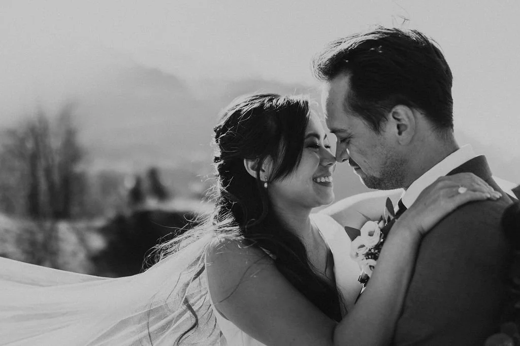 A black and white photo of a bride and groom, smiling and touching foreheads, embracing outdoors with a scenic background.
