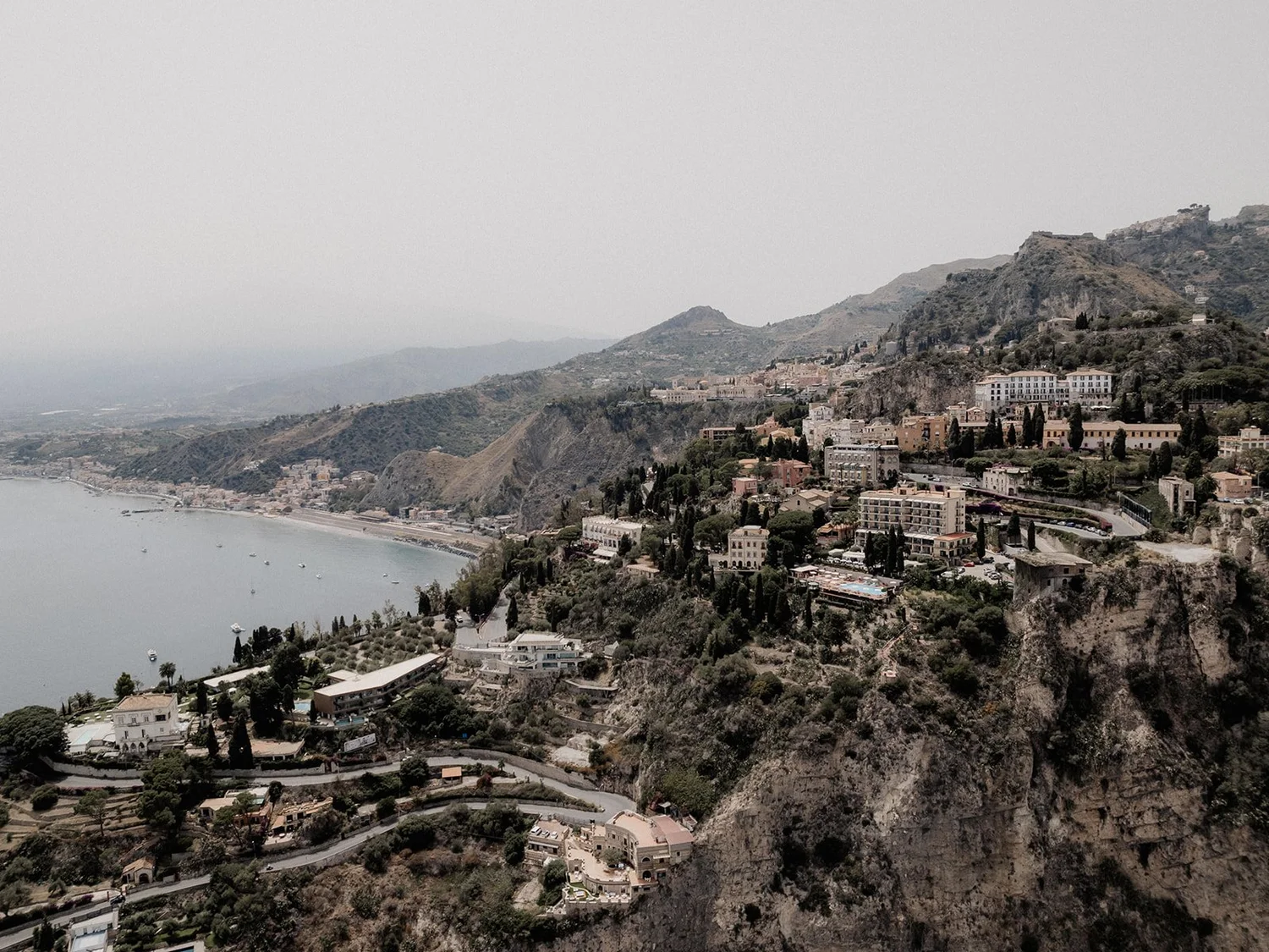 A coastal hillside town with numerous buildings and trees overlooking a bay with boats. Mountainous terrain surrounds the town.