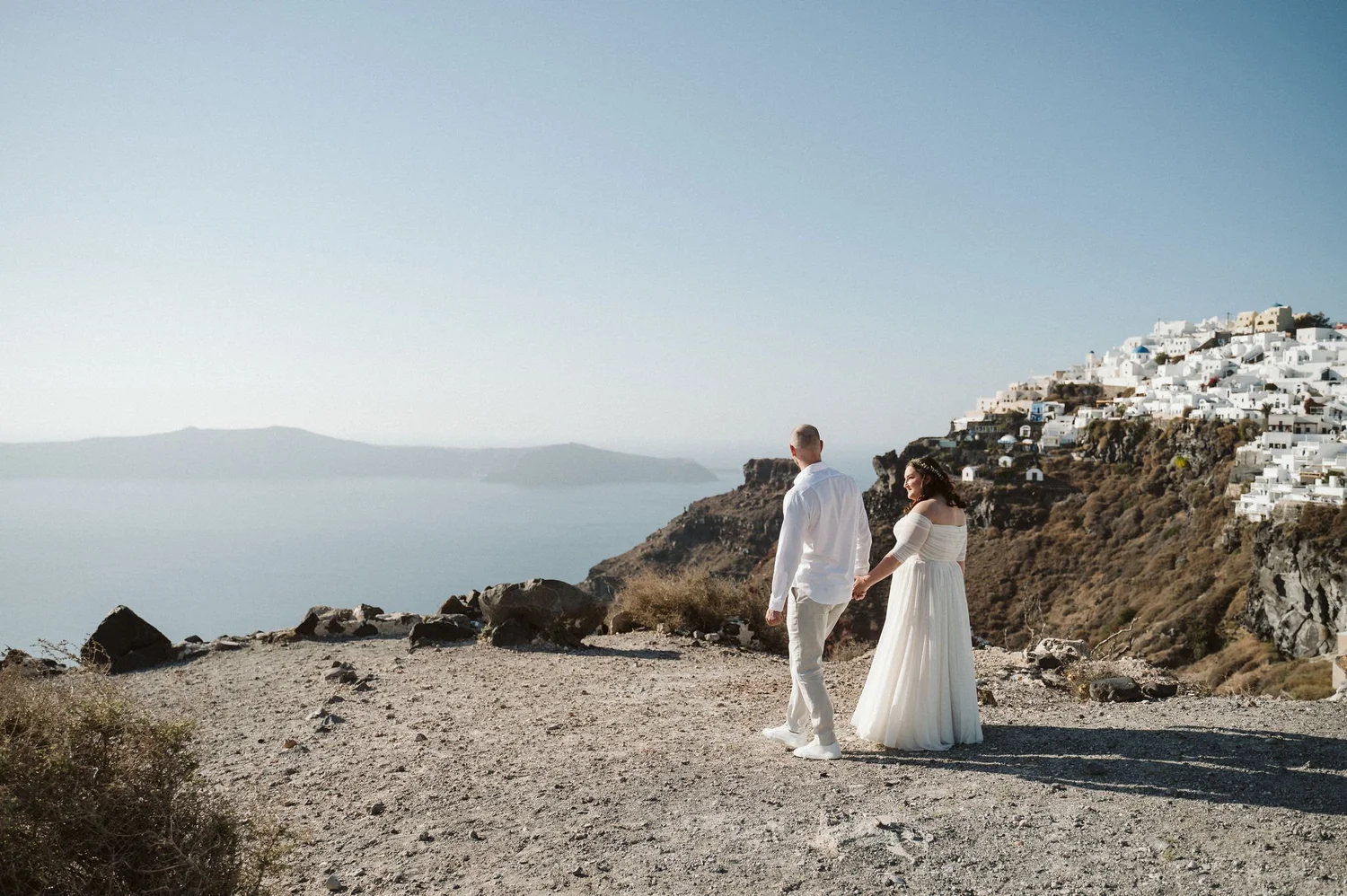 A couple dressed in white, holding hands and walking along a rocky path near the coast, with white buildings on a hillside in the background.