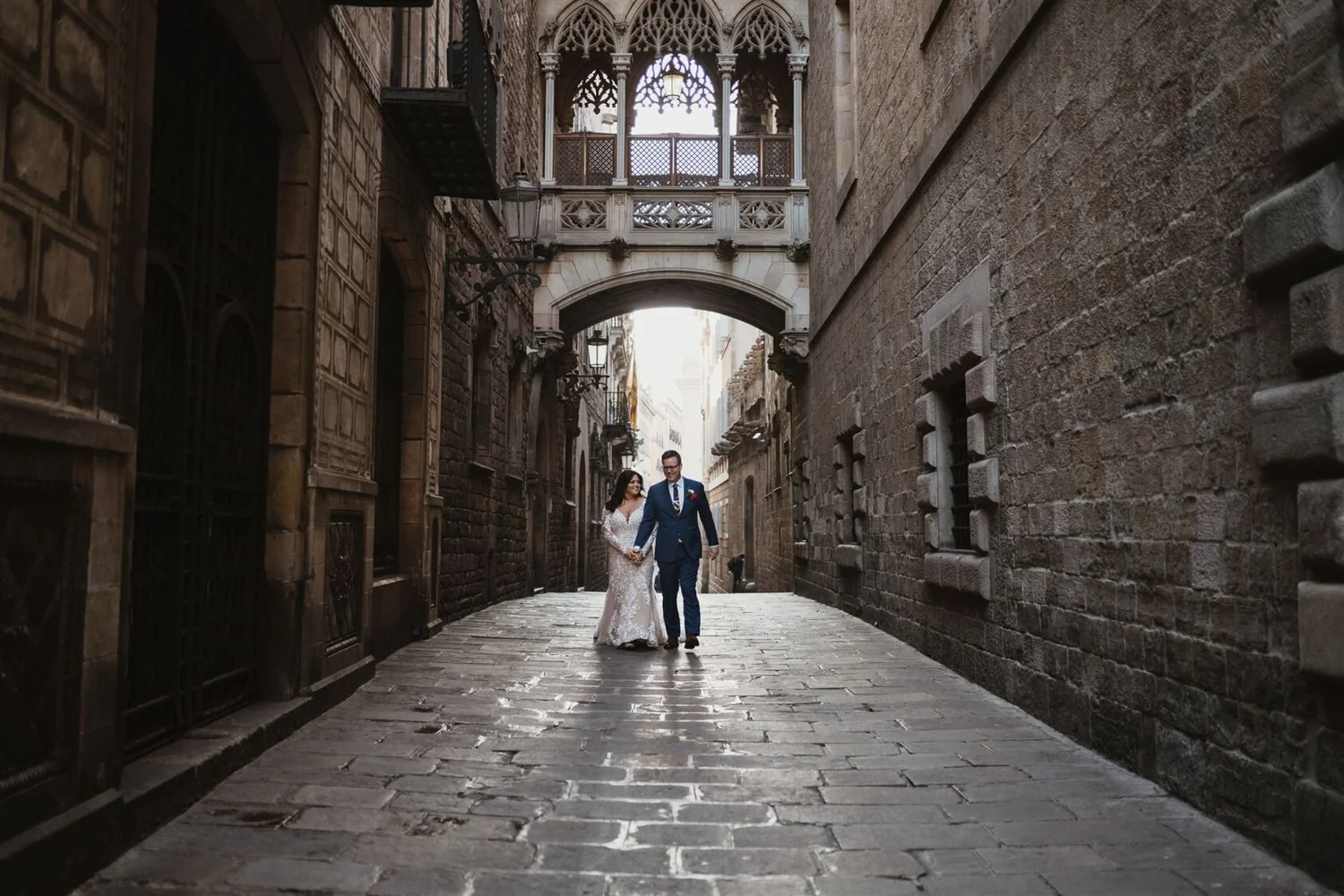 A bride and groom walking hand in hand through a narrow cobblestone street with old brick buildings and decorative architecture in the background.