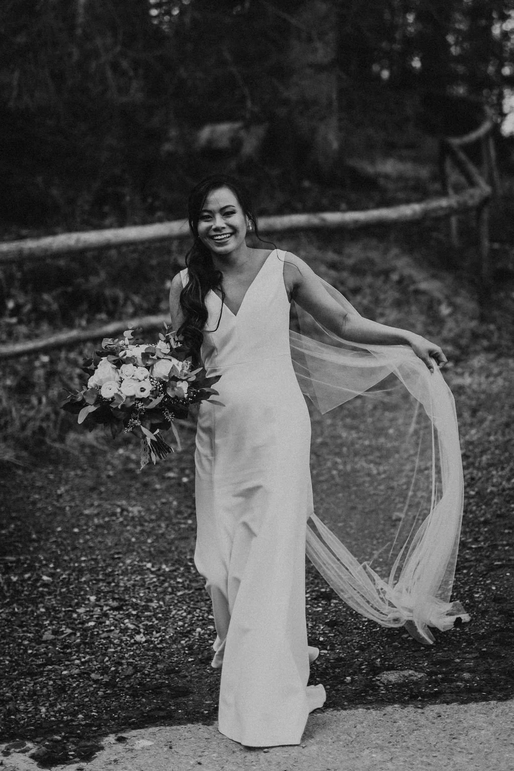 A smiling woman in a white wedding dress holding a bouquet of flowers, walking outdoors on a gravel path with trees in the background.