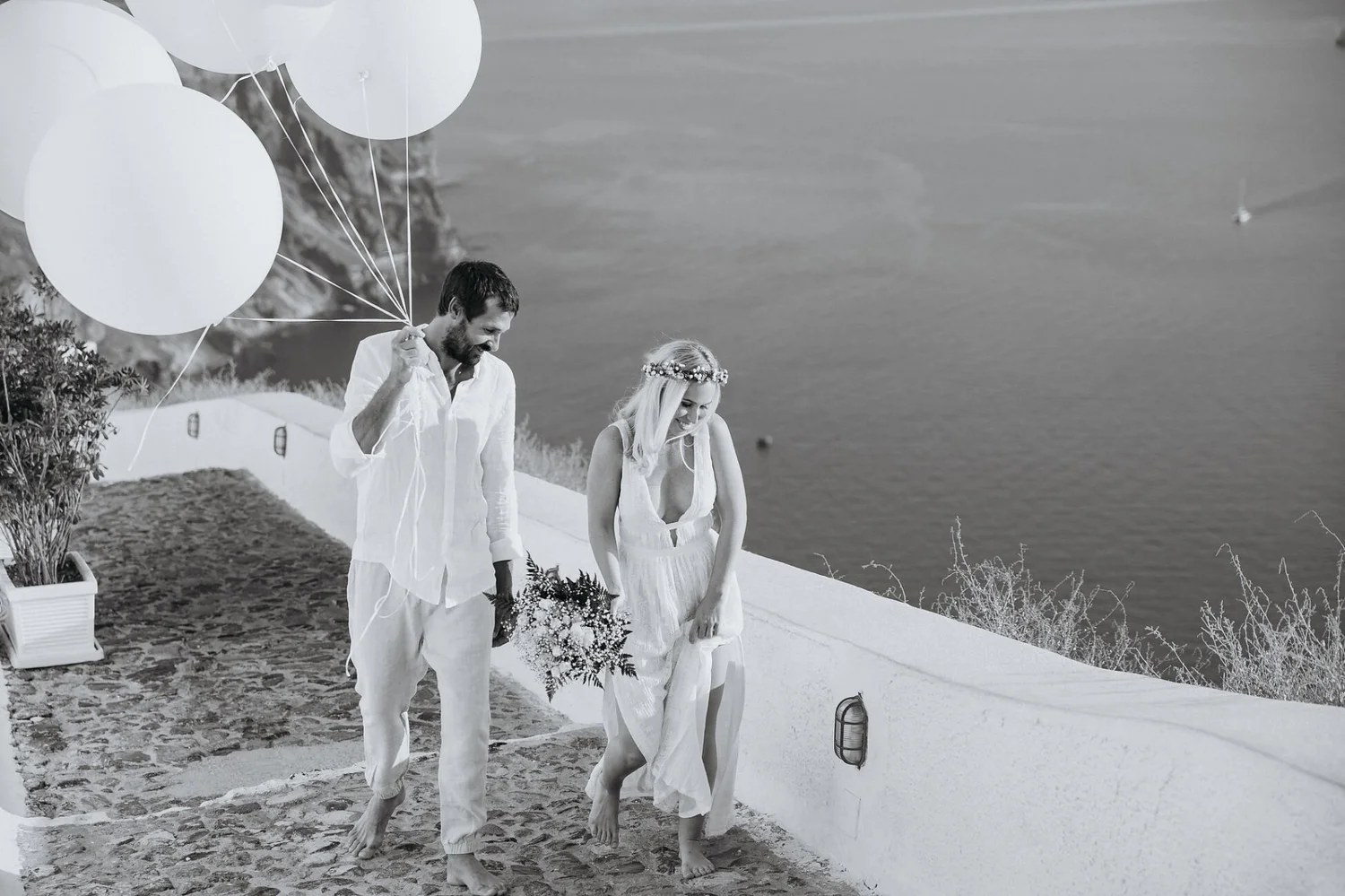A man and woman walking barefoot along a cobblestone path near water, holding hands. The man holds white balloons, and the woman has a flower crown and a bouquet of flowers. The scene appears to be a wedding or celebration near the coast.