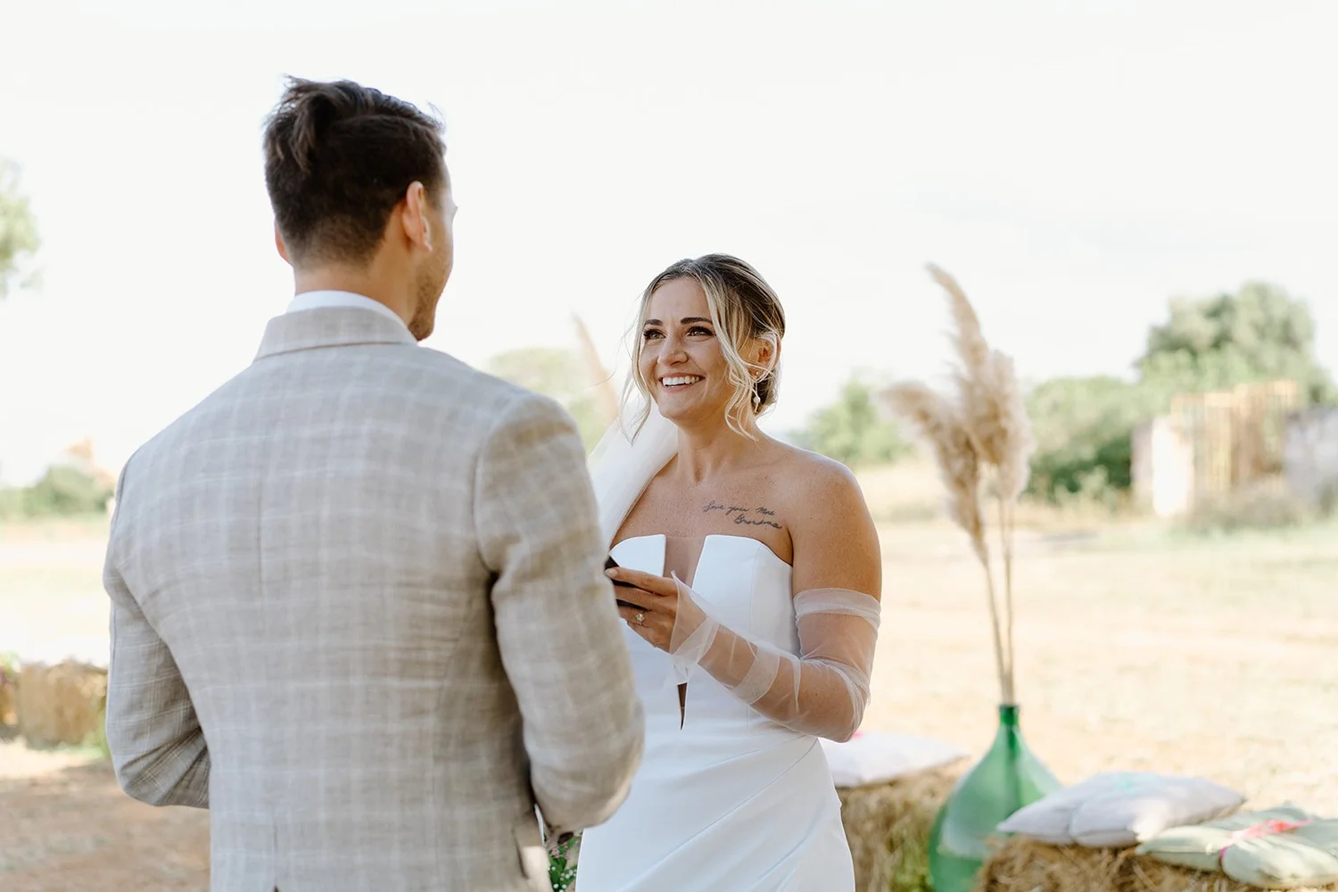A woman in a white wedding dress exchanging vows with a man in a light gray suit outdoors during daytime, with a scenic rural background.