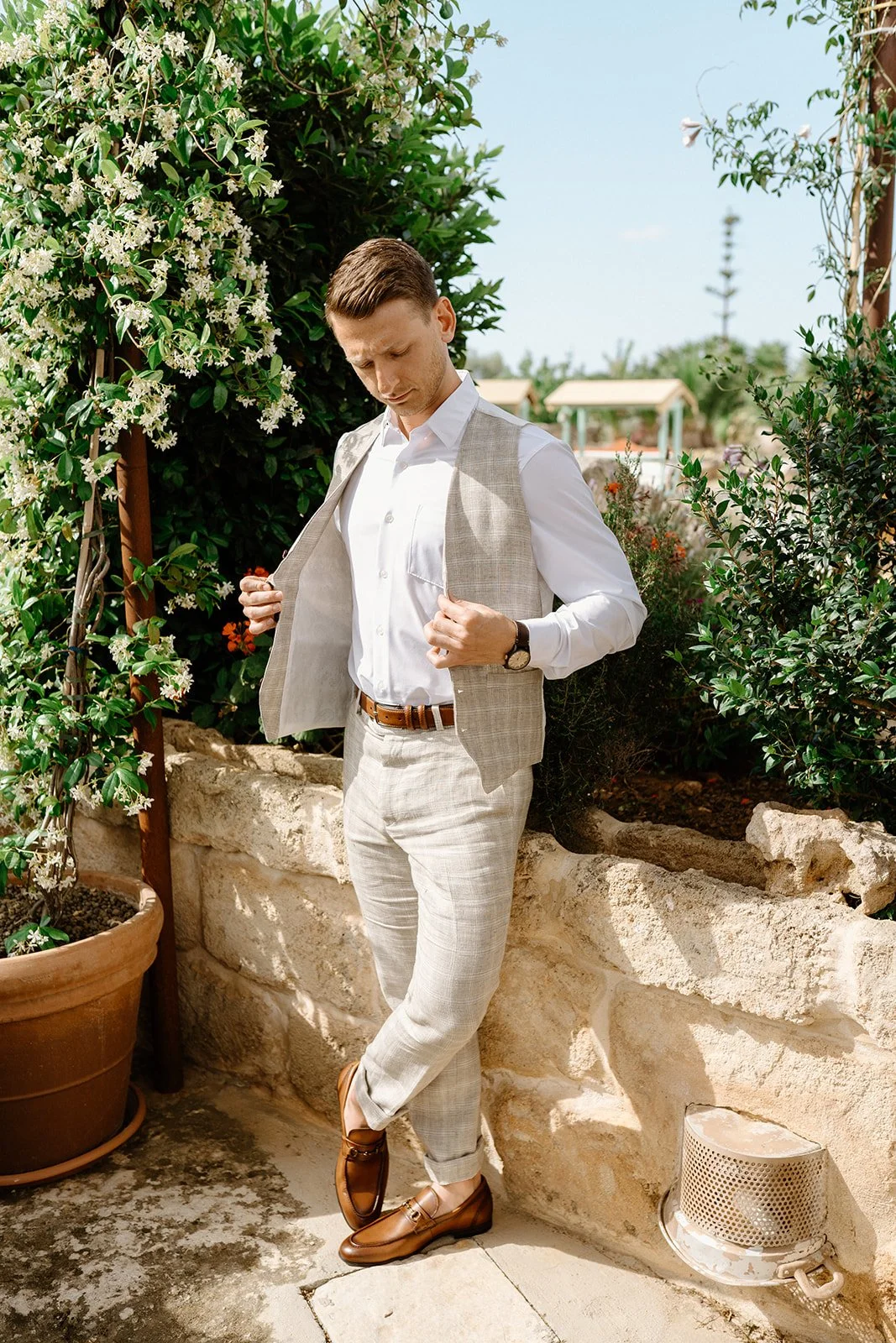 A man in light-colored pants, white shirt, and light gray vest standing outdoors near a stone wall and potted plants, adjusting his vest, with a garden and blue sky in the background.