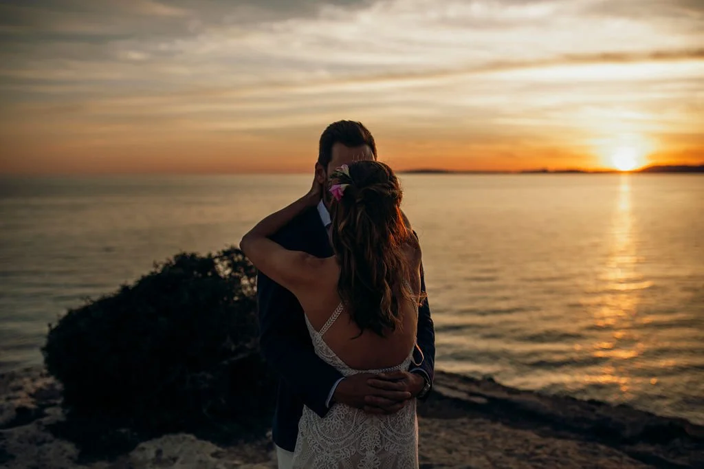 A couple embraces on a beach during sunset, with the ocean and a colorful sky in the background.