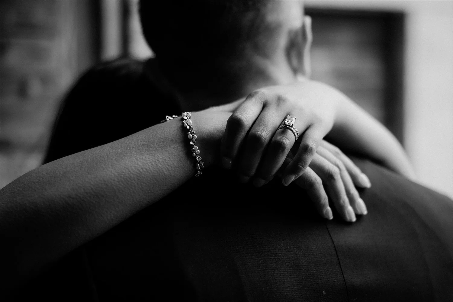 Close-up of two women embracing, with one woman resting her head on the other's shoulder. The image is in black and white, highlighting jewelry on the woman's hand and wrist, including a ring and a bracelet.