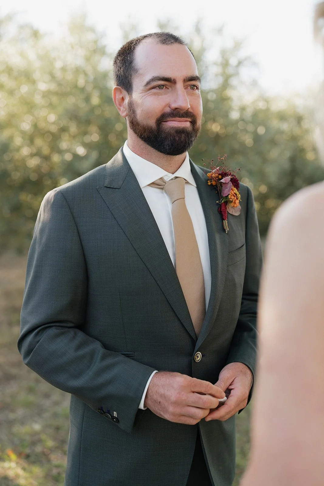 A groom with a beard and dark hair, wearing a gray suit with a cream tie and a floral boutonniere, standing outdoors during daylight with trees in the background, looking at someone off-camera.