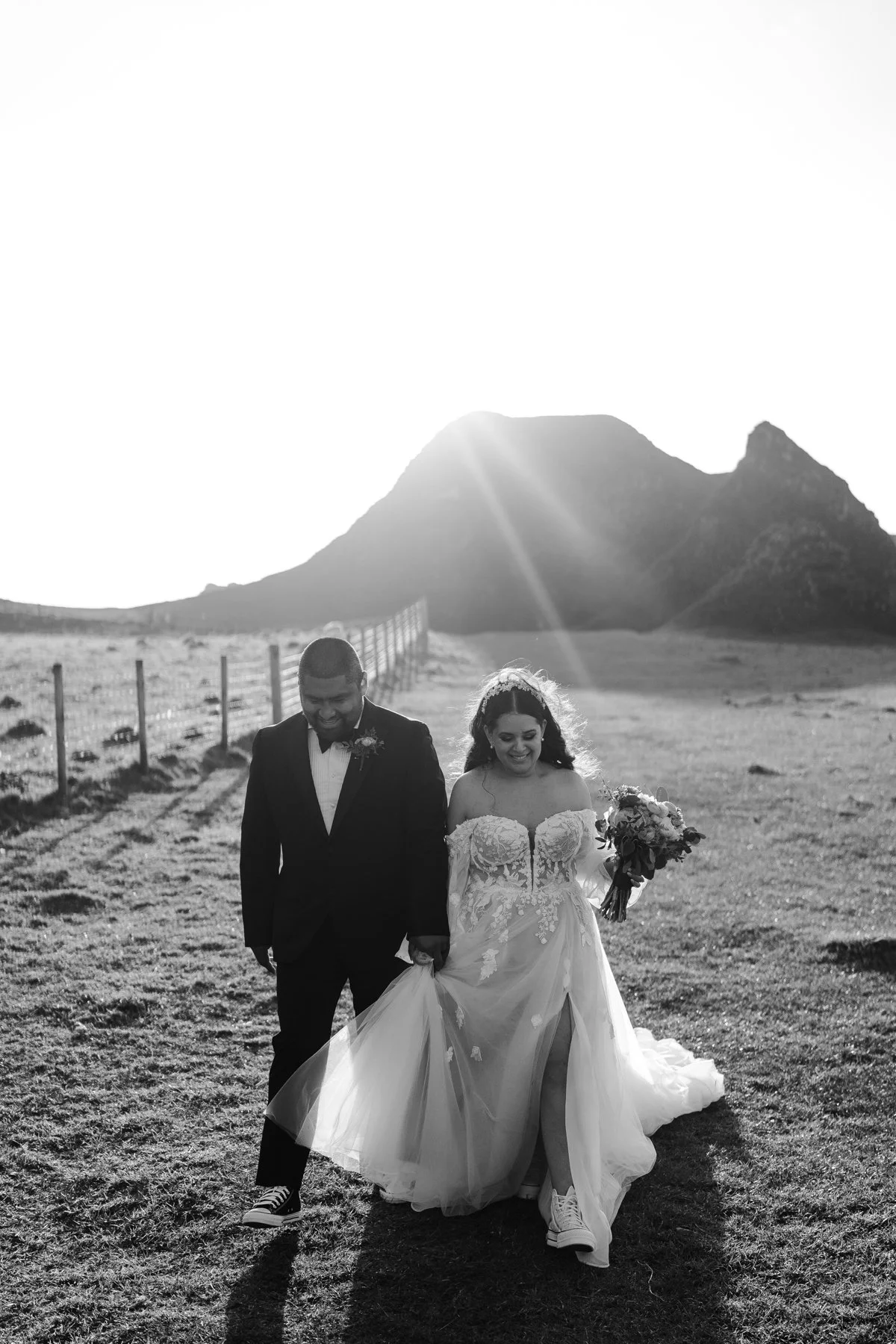 A black and white photograph of a bride and groom walking outdoors in a field at sunset. The bride is wearing a lace wedding gown, holding a bouquet, and wearing sneakers. The groom is in a tuxedo with a boutonniere and sneakers. They are smiling and