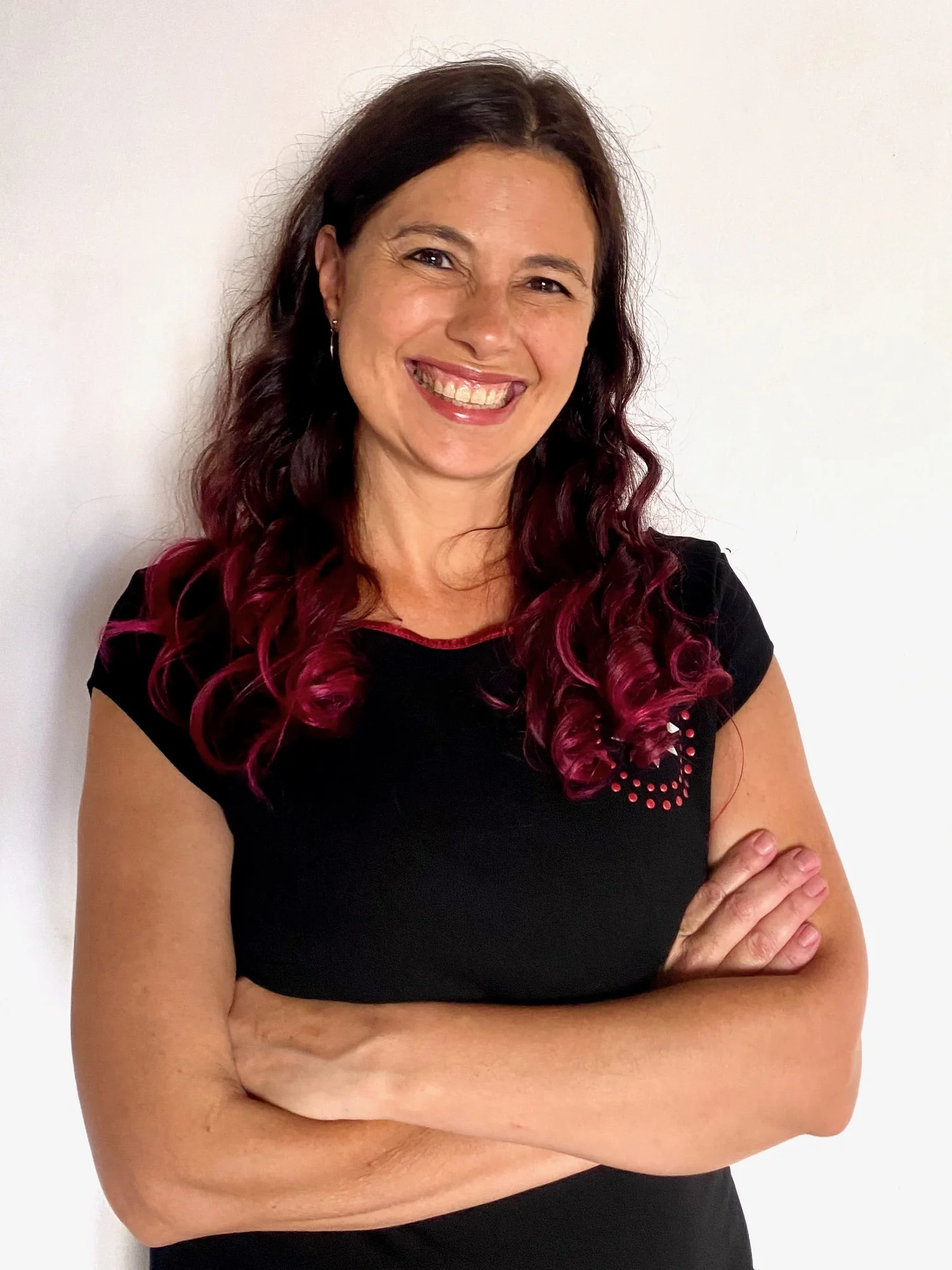 A woman with long, dark, and pink-colored curly hair smiling and crossing her arms, wearing a black t-shirt with pink accents.