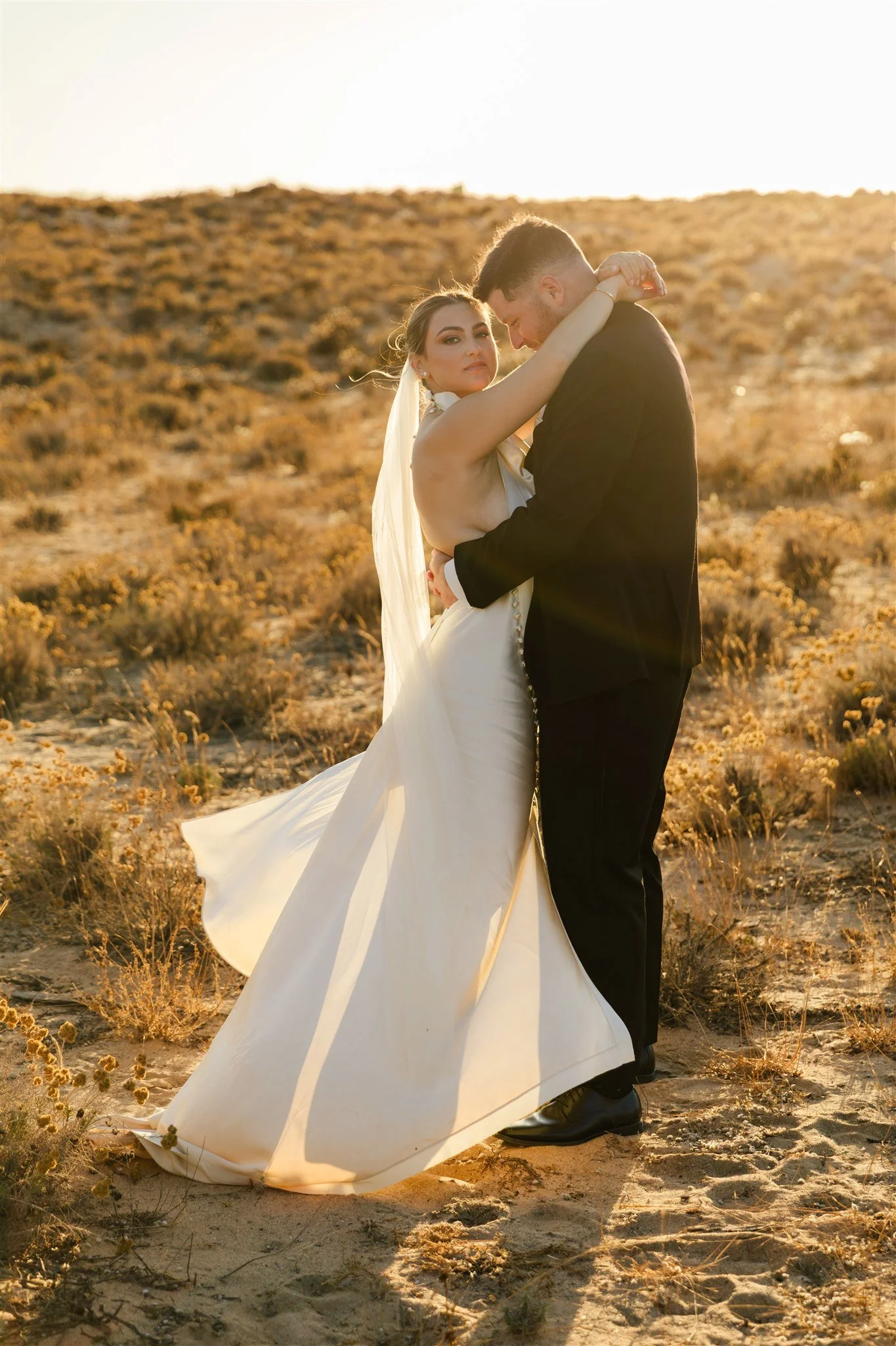 A bride and groom are embracing outdoors during sunset in a desert landscape, with the bride in a white wedding gown and the groom in a black suit.