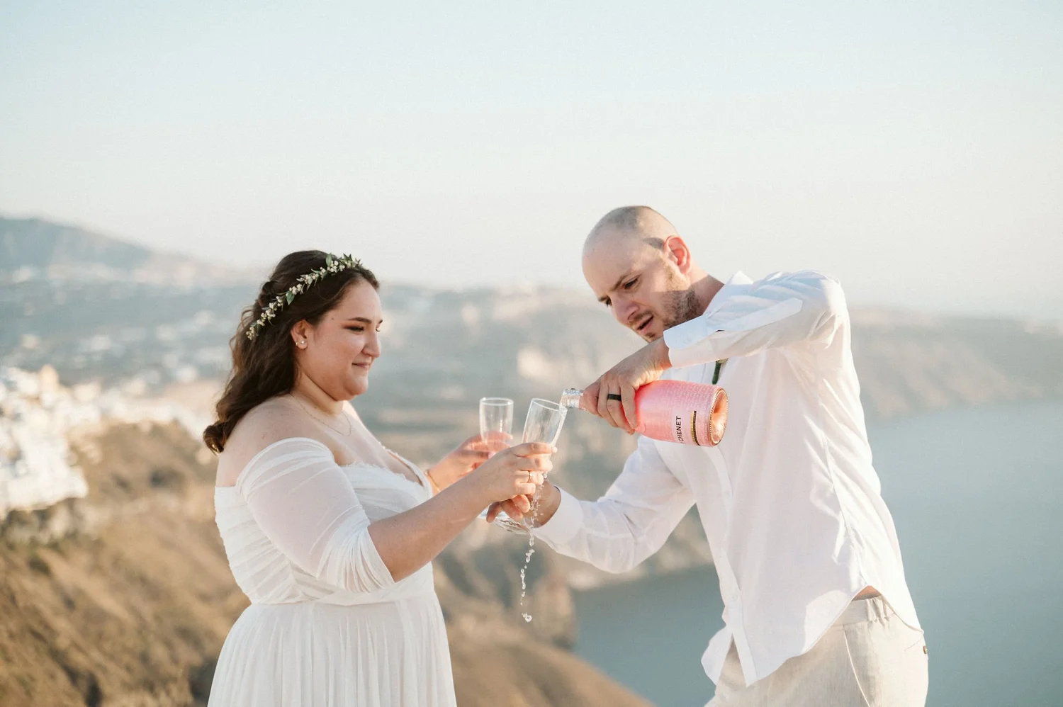 A couple celebrating a wedding on a scenic outdoor location, with the man pouring pink champagne into a fluted glass held by the woman, both wearing white attire.