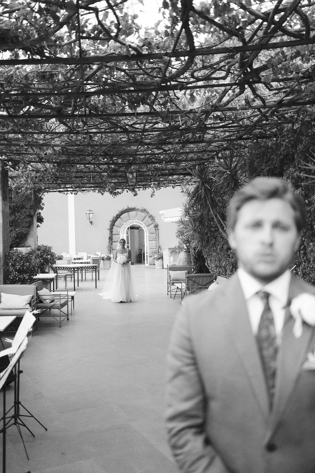 Black and white photo of a wedding scene with the bride in a gown holding a bouquet in the background, standing in an outdoor courtyard with chairs and tables. The groom, in a suit, is in the foreground and out of focus.
