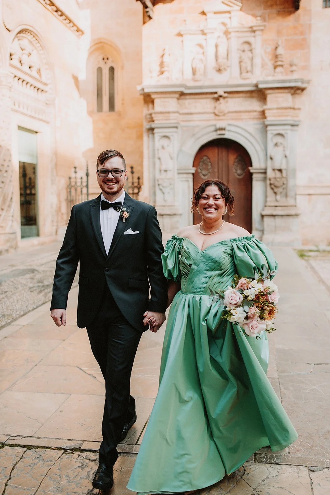 A smiling couple holding hands, walking outside a historic building with ornate stonework and a wooden arched door. The man is wearing a black tuxedo with a bow tie, and the woman is dressed in a green gown holding a bouquet of flowers.