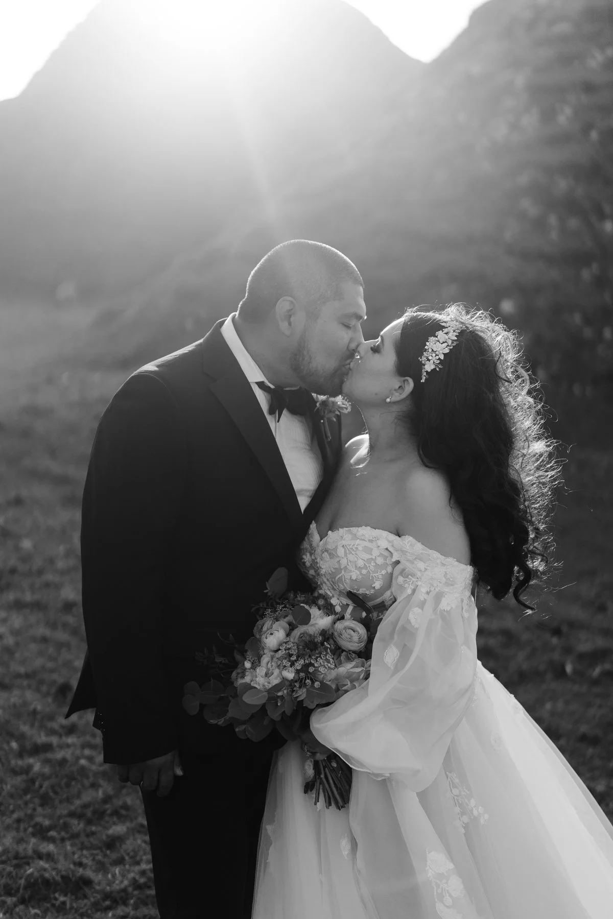 A black-and-white photo of a bride and groom kissing outdoors, with mountains and sunlight in the background. The bride is holding a bouquet and wearing a lace off-the-shoulder wedding dress, and the groom is in a tuxedo.